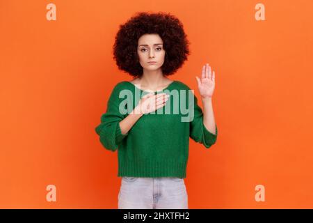 Ich verspreche. Seriöse Frau mit Afro-Frisur trägt grünen Pullover stehen Hand heben und schwören, Treue schwören, Treue versprechen. Innenaufnahme des Studios isoliert auf orangefarbenem Hintergrund. Stockfoto