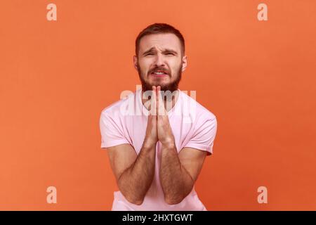 Porträt eines bärtigen Mannes, der im Gebet die Hände faltete, die Augen schloss und mit gott sprach, um Hilfe bat, Dankbarkeit ausdrückte und ein rosa T-Shirt trug. Innenaufnahme des Studios isoliert auf orangefarbenem Hintergrund. Stockfoto