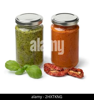 Glas mit rotem und grünem italienischen Pesto mit getrockneten süßen Tomaten und frischen Basilikumblättern vorne isoliert auf weißem Hintergrund Stockfoto