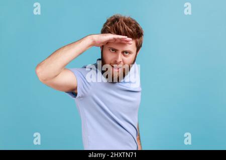 Porträt eines hübschen jungen erwachsenen bärtigen Mannes, der mit der Hand über dem Kopf weit weg schaut, sieht etwas angenehmes mit flirtendem Ausdruck. Innenaufnahme des Studios isoliert auf blauem Hintergrund. Stockfoto