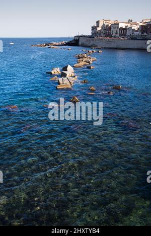 Felsenlinie im blauen Meer mit Gebäuden im Hintergrund in Ortigia, Sizilien, Italien Stockfoto
