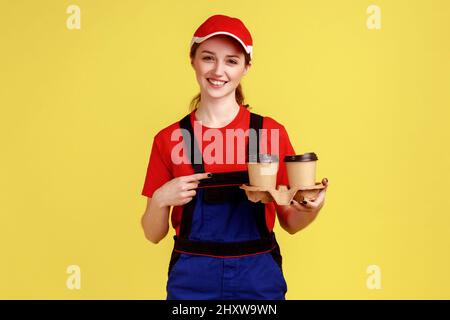 Porträt einer lächelnden, positiven Kurierin, die auf Kaffeetassen zeigt, lächelnd auf die Kamera blickt, Overalls und rote Mütze trägt. Innenaufnahme des Studios isoliert auf gelbem Hintergrund. Stockfoto