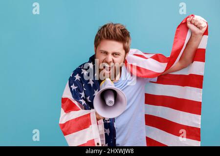 Porträt eines aufgeregten, selbstbewussten bärtigen Mannes, der auf Lautsprecher schreit und die Flagge der vereinigten Staaten von amerika hält, patriotischer Protest. Innenaufnahme des Studios isoliert auf blauem Hintergrund. Stockfoto