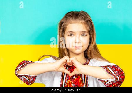 Ein Mädchen in ukrainischer Nationalkleidung, Vyschywanka, zeigt ein Herzzeichen als Zeichen der Liebe zur Ukraine, Nahaufnahme vor dem Hintergrund der Ukrainerin Stockfoto