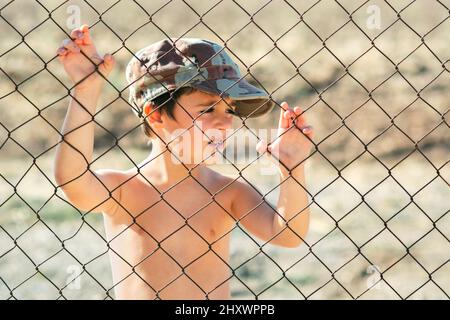 Trauriges Kind in einer Militärmütze hinter einem Zaun. Stop war Concept Stockfoto