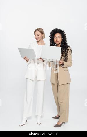 In voller Länge von stilvollen interracial Geschäftsfrauen mit Laptops auf grauem Hintergrund, Stockbild Stockfoto