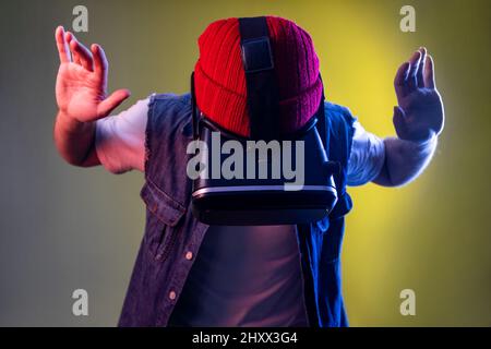 Gamer-Mann im Virtual-Reality-Headset, der mit erhobenem Arm steht, nach unten schaut, Spiel spielt oder fliegt, Beanie-Hut und Jeansweste trägt. Innenaufnahmen im Studio, isoliert auf farbigem Neonlicht-Hintergrund. Stockfoto