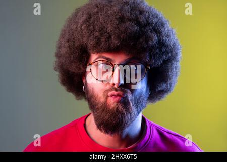 Porträt eines verrückten Hipster-Mannes mit Afro-Frisur, der mit gekreuzten Augen steht und mit einem lustigen Komiker-Gesicht aussieht, das ein rotes Sweatshirt trägt. Innenaufnahmen im Studio, isoliert auf farbigem Neonlicht-Hintergrund Stockfoto