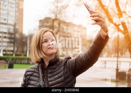 Porträt einer jungen glücklichen Frau, die in der Stadt Selfie macht. Sonne nach dem Regen. Selfie- und Videoanruf-Konzept. Stockfoto