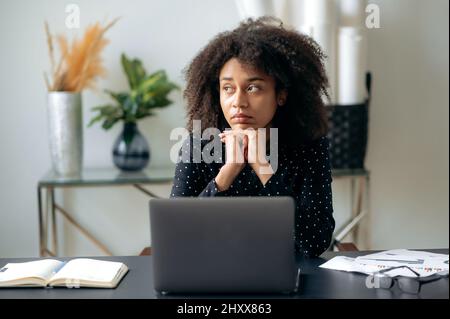 Müde afroamerikanische lockige Frau, Geschäftsfrau, Direktorin, Managerin, sitzt vor einem Laptop an ihrem Schreibtisch, ruht sich von der Computerarbeit aus, schaut zur Seite, träumt von Ruhe oder Urlaub Stockfoto