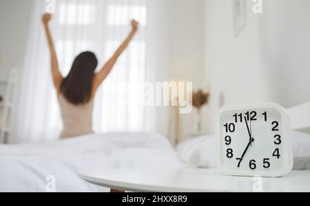 Nahaufnahme des Weckers vor dem Hintergrund, von dem sich die glückliche Frau, die auf dem Bett sitzt, nach dem Aufwachen streckt. Stockfoto