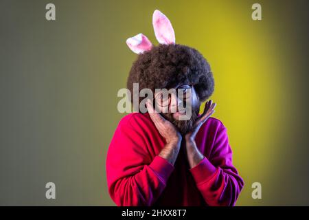 Porträt eines niedlichen charmanten Hipster-Mannes mit Afro-Frisur und Hasenohren, der mit einem Lächeln auf die Kamera blickt und ein rotes Sweatshirt trägt. Innenaufnahmen im Studio, isoliert auf farbigem Neonlicht-Hintergrund. Stockfoto