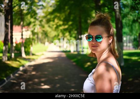 Nahaufnahme eines Porträts einer schönen glücklichen Frau in Sonnenbrillen, die an sonnigen Sommertagen im Park spazieren geht. Attraktive kaukasische Frau im Park. Stockfoto