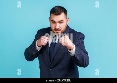 Porträt von herrisch strengen tragen offiziellen Stil Anzug klammernden Fäusten in Boxen Geste und tapfer Blick auf die Kamera, bereit zu schlagen. Innenaufnahme des Studios isoliert auf blauem Hintergrund. Stockfoto
