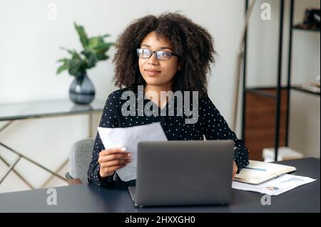 Attraktive, erfolgreiche junge Erwachsene dunkelhäutige Frau, Geschäftsführerin, Führungskraft oder Büroangestellte, sitzt im Büro, nutzt Laptop, arbeitet mit Dokumenten, Diagrammen, schaut weg, Analysiert die wirtschaftliche Dynamik Stockfoto