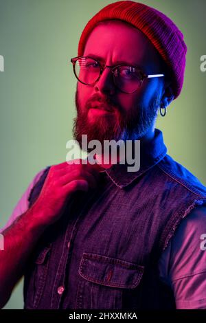 Porträt eines attraktiven bärtigen Hipsters in einer Brille, der die Kamera anschaut, die Hand am Kragen hält und einen roten Beanie-Hut trägt. Innenaufnahmen im Studio, isoliert auf farbigem Neonlicht-Hintergrund. Stockfoto