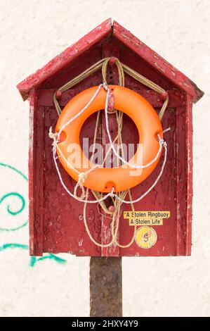 Eine vertikale Aufnahme eines orangefarbenen Rettungsring, der an einem kleinen roten Haus auf einem weißen Wandhintergrund hängt - das Konzept der Lebensleber Bedeutung Stockfoto
