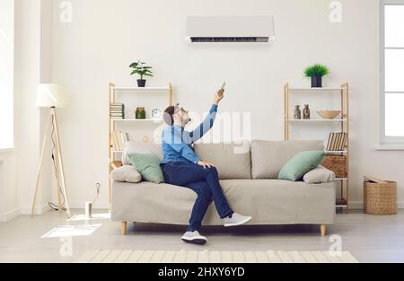 Der Mann, der zu Hause auf der Couch sitzt, schaltet die Klimaanlage per Fernbedienung Stockfoto