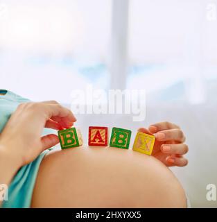 Die größte Freude ist auf dem Weg. Nahaufnahme einer Schwangeren, die zu Hause mit hölzernen Babyblöcken auf ihrem Bauch liegt. Stockfoto