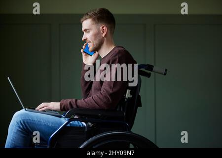 Mann im Rollstuhl, der von zu Hause aus mit Laptop und Mobiltelefon arbeitet Stockfoto
