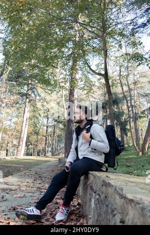 Ein Mann, der in der Ecke des Waldes sitzt. Bäume hinter ihm. Mann, der weit weg schaut. Sportlicher Kerl im Wald. Fahrt zwischen Bäumen. Stockfoto