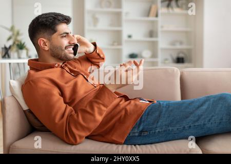 Fröhlicher arabischer Mann, der auf dem Handy spricht und auf der Couch liegt Stockfoto