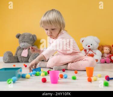 Ein zwei Jahre altes Mädchen sitzt auf dem Boden Spielzimmer vor dem Hintergrund einer gelben Wand und legt Kugeln in bunten Tassen nach Farbe Stockfoto