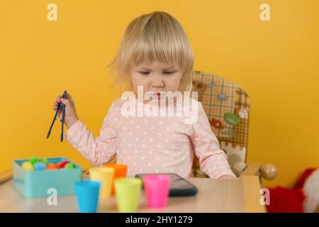 Ein zweijähriges Mädchen in rosa Kleidung an einem Tisch gegen die gelbe Wand legt Kugeln in bunten Tassen nach Farben aus und schaut in das Smartphone Stockfoto