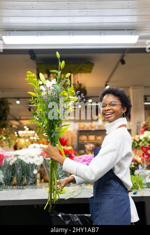 Positive afroamerikanische weibliche Floristin mit Blumenstrauß mit grünen Ästen, die während der Arbeit in einem Floristikgeschäft die Kamera anschaut Stockfoto