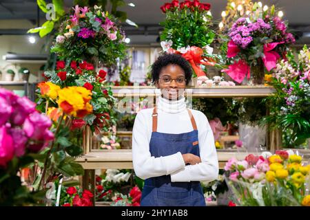 Lächelnde afroamerikanische Floristin in Schürze, die in einem Blumengeschäft zwischen bunten Blumensträußen steht und die Kamera anschaut Stockfoto