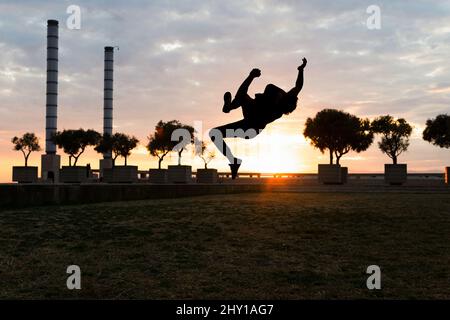 Voller Körper von unkenntlich männlichen Silhouette springen über dem Boden mit erhobenen Armen während Parkour Trick bei Sonnenuntergang Stockfoto