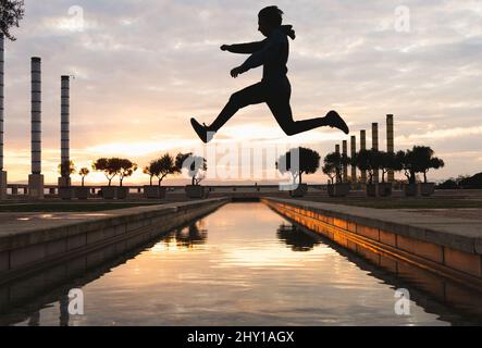 Voller Körper von unkenntlichen männlichen Silhouette springen über See, während Parkour Trick bei Sonnenuntergang Stockfoto