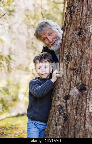 Positiver älterer Großvater mit grauem Haar und Bart, mit einem kleinen lächelnden Jungen, der sich hinter einem Baum versteckt und vor verschwommenem Hintergrund die Kamera im Wald anschaut Stockfoto