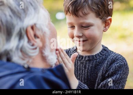 Fröhlicher kleiner Enkel in warmem Pullover, der den grauen Bart eines lächelnden Großvaters mit geschlossenen Augen auf verschwommenem Hintergrund berührt Stockfoto