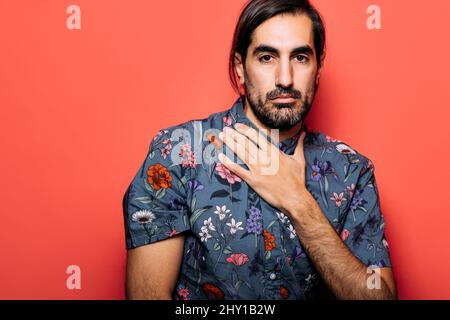 Seriöser Mann mit schwarzem Haar und Bart im farbenfrohen Hemd, der die Kamera anschaut, während er auf rotem Hintergrund im hellen Studio steht Stockfoto