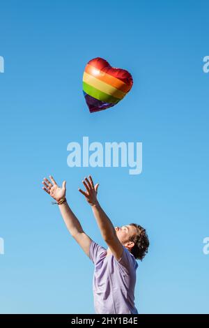 Seitenansicht eines transgender männlichen Mannes mit erhobenen Armen, der einen herzförmigen Ballon mit einem LGBT-Symbol in Regenbogenfarben in der Luft gegen den blauen Himmel auslöst Stockfoto