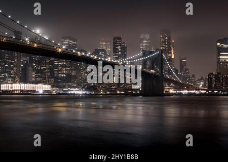 Beleuchtete Brücke über den ruhigen Fluss mit modernen Hochhäusern und leuchtenden Lichtern in New York City bei Nacht Stockfoto