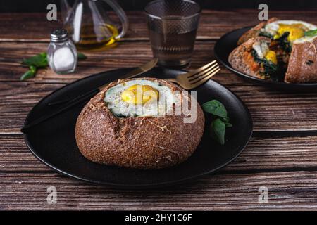 Leckeres rundes Brot mit Spiegelei auf schwarzem Teller auf Holztisch mit Besteck in hellem Restaurant serviert Stockfoto
