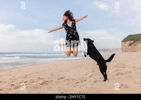 Ein ganzer Körper aktiver weiblicher Reisender, die mit ihrem schwarzen Hund und dem Meer auf der Sandküste springt, während der Reise in einem tropischen Resort Stockfoto