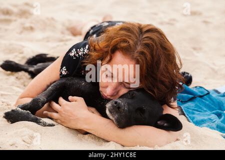 Schläfrige Besitzerin mit niedlichem gehorsamen schwarzen Hund, der am Sandstrand schläft, während er sich in den Sommerferien im tropischen Resort ausruhen kann Stockfoto