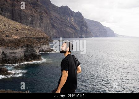 Seitenansicht nachdenklicher männlicher Tourist, der die Küste von rauer Küste unter düsterem Himmel während der Reise auf Teneriffa bewundert Stockfoto