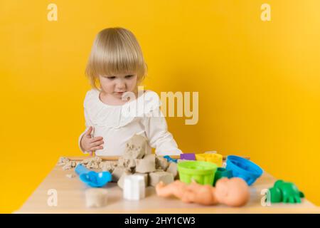 Ein schönes Mädchen in einem weißen Kleid steht neben einem Tisch mit kinetischem Sand, Plastikformen und anderem Spielzeug. Selektiver Fokus Stockfoto