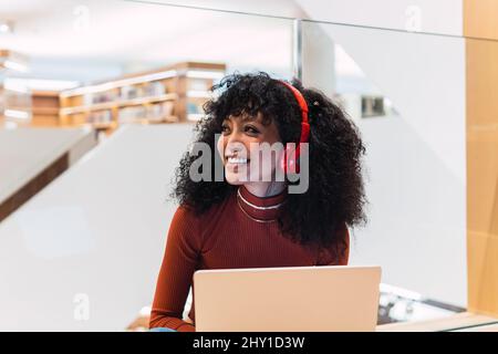 Schwarze, lockige Frau mit Kopfhörern, die lächelnd mit einem Laptop in der modernen Bibliothek sitzt Stockfoto