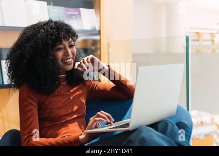 Zufriedene afroamerikanische Frau mit Afro-Frisur, die in einem Sessel in der Bibliothek sitzt und einen Laptop benutzt, während sie an einem Projekt arbeitet Stockfoto