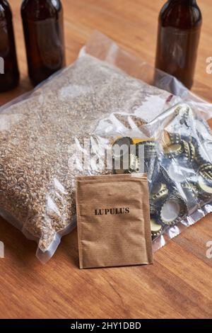 Pakete mit Gerstenkörnern und Metallkappen in der Nähe von Hopfen für das Brauen von hausgemachtem Bier auf dem Tisch mit Glasflaschen im Zimmer platziert Stockfoto