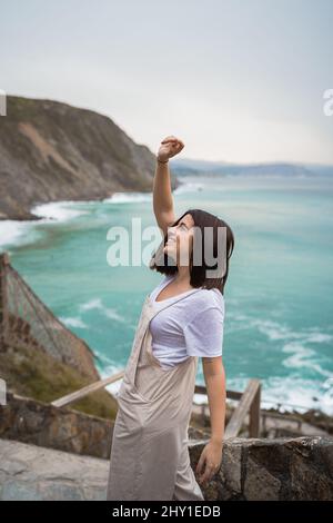 Attraktive Weibchen im Ganzen wegschauen, während sie auf felsiger Küste in der Nähe von plätscherndem Meer während der Reise in Küstengebiet stehen Stockfoto