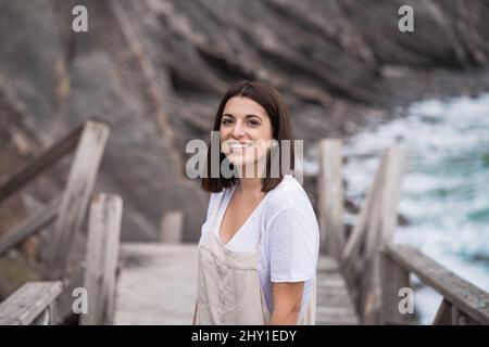 Charmante junge Frau im Ganzen Blick auf die Kamera mit einem Lächeln, während auf Holztreppe in der Natur gegen Berghang stehen Stockfoto