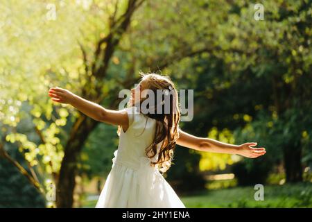 Glückliches junges Mädchen in Erstkommunion Kleid lächelnd und Blick auf weg, während im grünen Park an sonnigen Tag tanzen Stockfoto