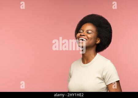 Fröhliche afrikanische Frau lacht über rosa Wand. Entzückendes junges Mädchen lächelt mit geschlossenen Augen Stockfoto