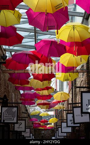 Farbenfrohe, gelbe und rote Regenschirme hängen über einer Gasse voller Boutiquen im Camden Market im Norden Londons. Stockfoto